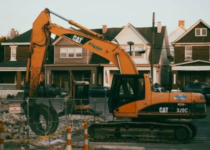 Excavator working in a suburban neighborhood construction site, with residential houses in the background.