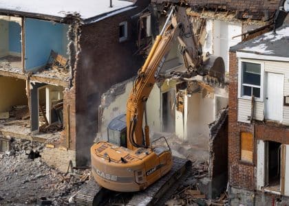 An excavator demolishes an old brick building at a construction site, creating debris and dust.